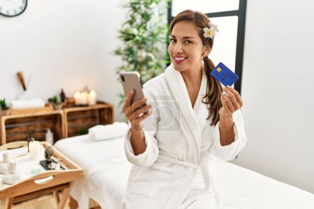 Photo for Young latin woman wearing bathrobe using smartphone and credit card - Royalty Free Image