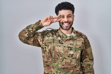 Photo for Arab man wearing camouflage army uniform doing peace symbol with fingers over face, smiling cheerful showing victory - Royalty Free Image