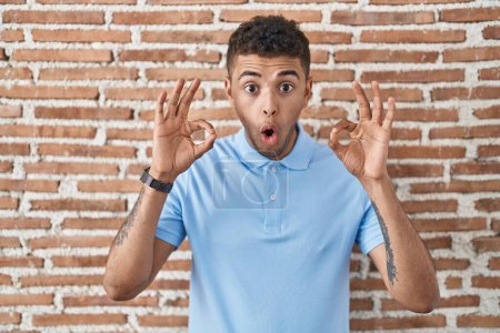 Téléchargez les photos : Jeune homme brésilien debout au-dessus du mur de briques regardant surpris et choqué faisant ok symbole d'approbation avec les doigts. expression folle - en image libre de droit