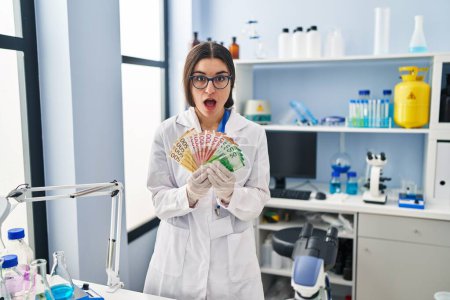 Photo for Young hispanic woman working at scientist laboratory holding money banknotes afraid and shocked with surprise and amazed expression, fear and excited face. - Royalty Free Image