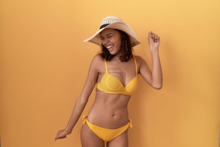Téléchargez les photos : Jeune femme hispanique portant un bikini et un chapeau d'été dansant heureux et joyeux, souriant, décontracté et confiant en écoutant de la musique - en image libre de droit