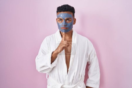 Téléchargez les photos : Jeune homme hispanique portant un masque de beauté et peignoir de bain pensée concentré sur le doute avec le doigt sur le menton et la recherche jusqu'à se demander - en image libre de droit