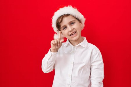 Foto de Pequeño chico caucásico con sombrero de navidad sobre fondo rojo sonriendo con una idea o pregunta apuntando con el dedo hacia arriba con la cara feliz, número uno - Imagen libre de derechos