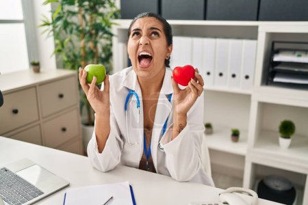 Téléchargez les photos : Jeune femme hispanique travaillant à la clinique diététiste tenant pomme verte en colère et folle hurlant frustré et furieux, criant avec colère levant les yeux. - en image libre de droit