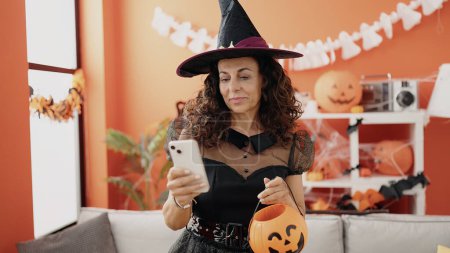 Téléchargez les photos : Moyen Age femme hispanique en utilisant smartphone ayant fête d'Halloween à la maison - en image libre de droit