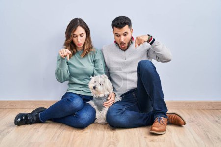 Photo for Young hispanic couple sitting on the floor with dog pointing down with fingers showing advertisement, surprised face and open mouth - Royalty Free Image