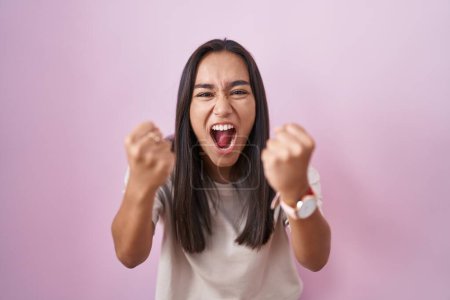 Photo for Young hispanic woman standing over pink background angry and mad raising fists frustrated and furious while shouting with anger. rage and aggressive concept. - Royalty Free Image