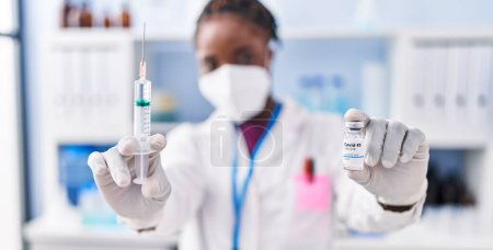 Photo for African american woman wearing scientist uniform and medical mask holding covid-19 vaccine at laboratory - Royalty Free Image