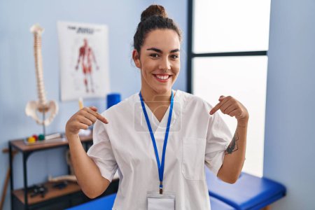 Photo for Young hispanic woman working at rehabilitation clinic looking confident with smile on face, pointing oneself with fingers proud and happy. - Royalty Free Image