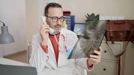 Foto de Médico de mediana edad hablando por teléfono mirando rayos X en la clínica - Imagen libre de derechos
