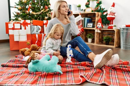 Foto de Madre e hija jugando con juguetes y hablando en el teléfono inteligente sentado junto al árbol de Navidad en casa - Imagen libre de derechos