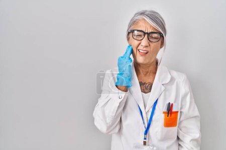 Foto de Mujer de mediana edad con pelo gris vistiendo bata científica tocando la boca con la mano con expresión dolorosa debido a dolor de muelas o enfermedad dental en los dientes. dentista - Imagen libre de derechos