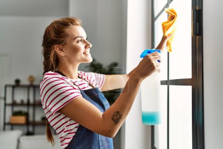 Téléchargez les photos : Jeune femme souriant confiant fenêtre de nettoyage à la maison - en image libre de droit