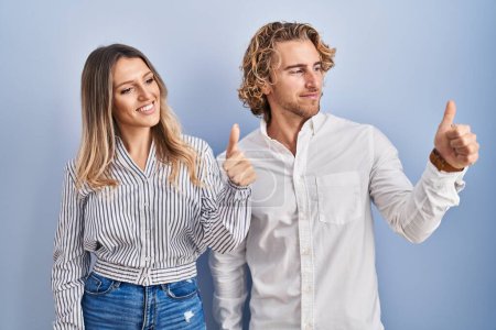 Téléchargez les photos : Young couple standing over blue background looking proud, smiling doing thumbs up gesture to the side - en image libre de droit