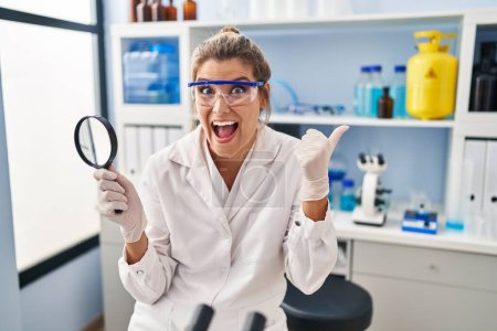 Foto de Young woman working at scientist laboratory holding magnifying glass pointing thumb up to the side smiling happy with open mouth - Imagen libre de derechos