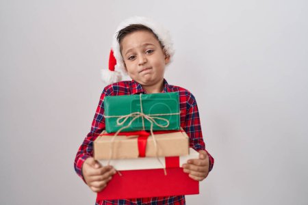 Photo for Little hispanic boy wearing christmas hat holding presents clueless and confused expression. doubt concept. - Royalty Free Image