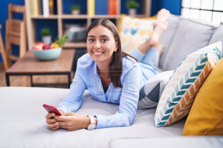 Foto de Mujer hispana hermosa joven usando teléfono inteligente tumbado en el sofá en casa - Imagen libre de derechos