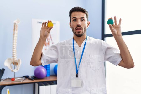 Foto de Joven fisioterapeuta hispano hombre sosteniendo bolas de fuerza para entrenar los músculos de la mano en la cara de choque, con aspecto escéptico y sarcástico, sorprendido con la boca abierta - Imagen libre de derechos