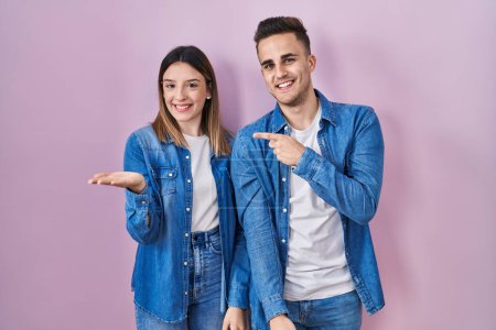 Téléchargez les photos : Jeune couple hispanique debout sur fond rose étonné et souriant à la caméra tout en présentant avec la main et pointant du doigt. - en image libre de droit