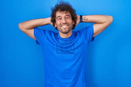 Photo for Hispanic young man standing over blue background relaxing and stretching, arms and hands behind head and neck smiling happy - Royalty Free Image