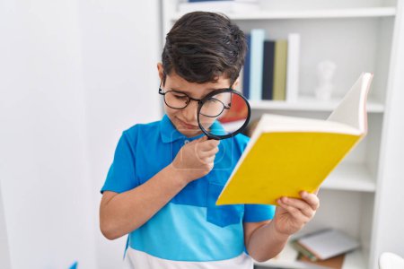 Photo for Adorable hispanic boy student reading book using magnifying glass at library school - Royalty Free Image