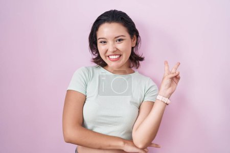 Photo for Hispanic young woman standing over pink background smiling with happy face winking at the camera doing victory sign with fingers. number two. - Royalty Free Image