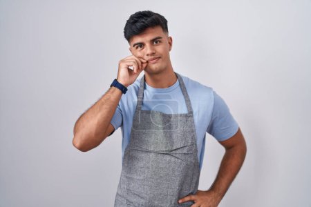 Photo for Hispanic young man wearing apron over white background mouth and lips shut as zip with fingers. secret and silent, taboo talking - Royalty Free Image