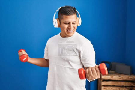 Téléchargez les photos : Jeune homme latin écoutant de la musique à l'aide d'haltères de formation au centre sportif - en image libre de droit