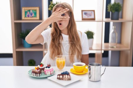 Foto de Mujer caucásica joven comiendo pasteles t para el desayuno asomándose en shock cubriendo la cara y los ojos con la mano, mirando a través de los dedos con expresión avergonzada. - Imagen libre de derechos