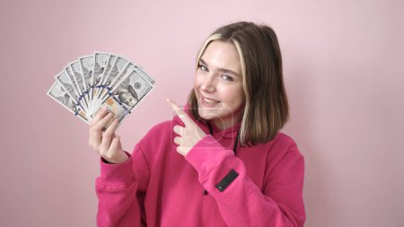 Foto de Joven mujer rubia sonriendo confiada señalando con el dedo a los dólares sobre el fondo rosa aislado - Imagen libre de derechos