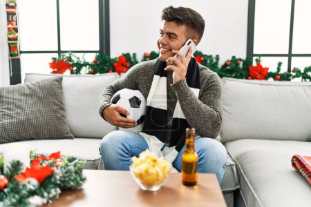 Téléchargez les photos : Jeune homme hispanique soutenant le jeu de football parler sur le smartphone assis sur le canapé par décor de Noël à la maison - en image libre de droit