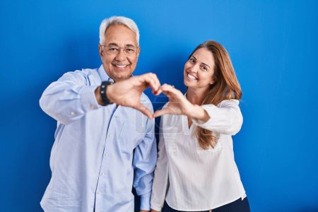 Téléchargez les photos : Couple hispanique du Moyen Age debout sur fond bleu souriant dans l'amour faisant forme de symbole de coeur avec les mains. concept romantique. - en image libre de droit