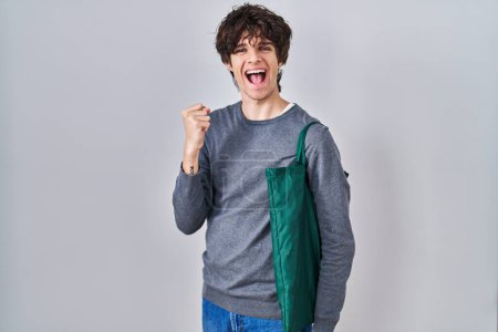 Téléchargez les photos : Young man wearing reusable bag screaming proud, celebrating victory and success very excited with raised arms - en image libre de droit
