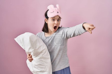 Foto de Woman with down syndrome wearing sleeping mask hugging pillow pointing with finger surprised ahead, open mouth amazed expression, something on the front - Imagen libre de derechos