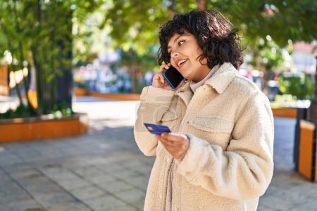 Téléchargez les photos : Jeune belle femme hispanique parlant sur smartphone tenant la carte de crédit au parc - en image libre de droit
