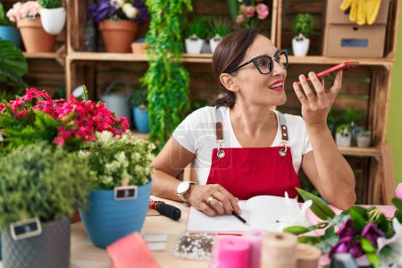 Foto de Young beautiful hispanic woman florist talking on smartphone sitting on table at flower shop - Imagen libre de derechos