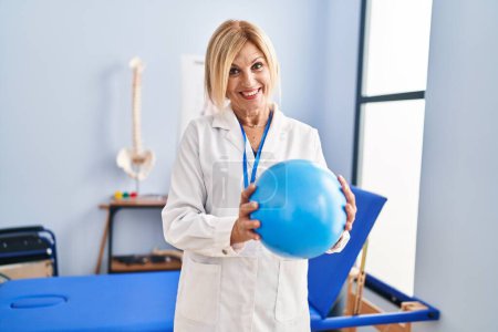 Téléchargez les photos : Femme blonde d'âge moyen portant un uniforme de physiothérapeute tenant le ballon à la clinique de physiothérapie - en image libre de droit