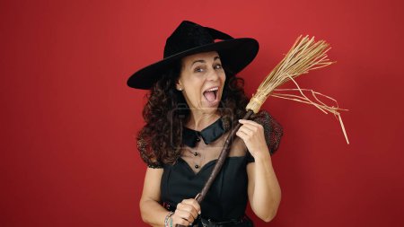 Téléchargez les photos : Middle age hispanic woman wearing witch costume holding broom over isolated red background - en image libre de droit