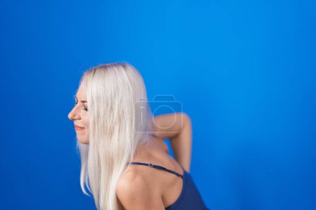 Foto de Mujer caucásica de pie sobre fondo azul sufriendo de dolor de espalda, tocando la espalda con la mano, dolor muscular - Imagen libre de derechos