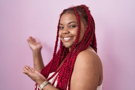 Téléchargez les photos : Femme afro-américaine aux cheveux tressés debout sur fond rose invitant à entrer souriant naturel à la main ouverte - en image libre de droit