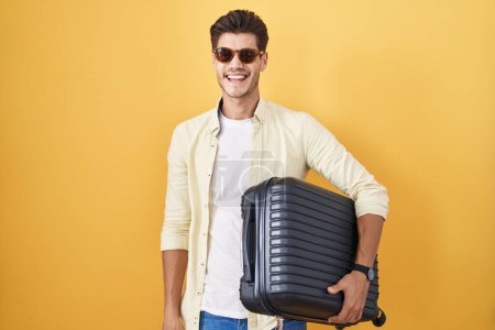 Foto de Joven hombre hispano sosteniendo maleta yendo de vacaciones de verano guiñando el ojo mirando a la cámara con expresión sexy, cara alegre y feliz. - Imagen libre de derechos