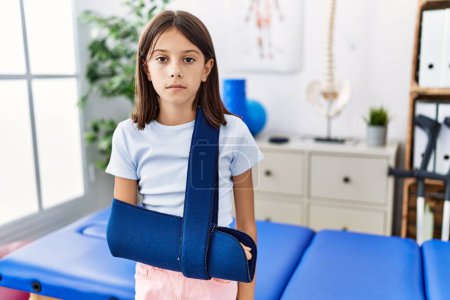 Foto de Young hispanic girl wearing arm on sling at rehabilitation clinic thinking attitude and sober expression looking self confident - Imagen libre de derechos