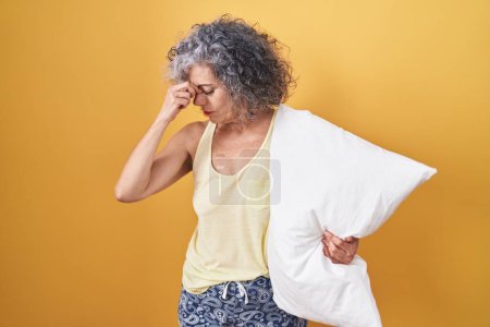 Foto de Middle age woman with grey hair wearing pijama hugging pillow tired rubbing nose and eyes feeling fatigue and headache. stress and frustration concept. - Imagen libre de derechos