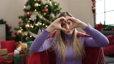 Photo for Young blonde woman doing heart gesture sitting on sofa by christmas tree at home - Royalty Free Image