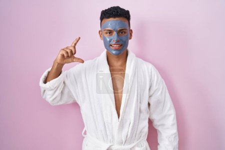 Foto de Young hispanic man wearing beauty face mask and bath robe smiling and confident gesturing with hand doing small size sign with fingers looking and the camera. measure concept. - Imagen libre de derechos