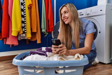 Photo for Young blonde woman using smartphone and washing clothes at laundry room - Royalty Free Image