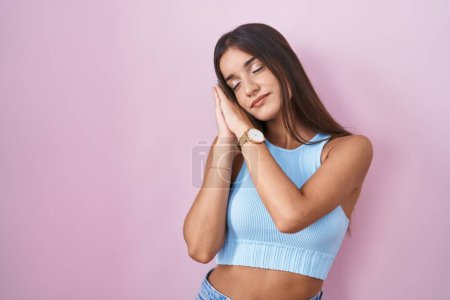 Téléchargez les photos : Jeune femme brune debout sur fond rose endormi fatigué rêvant et posant les mains ensemble tout en souriant les yeux fermés. - en image libre de droit