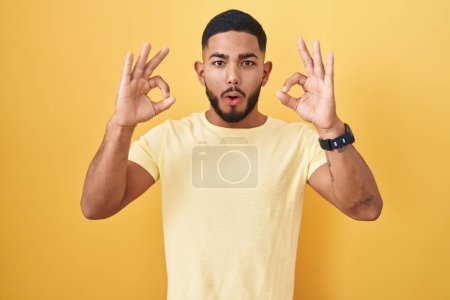 Photo for Young hispanic man standing over yellow background looking surprised and shocked doing ok approval symbol with fingers. crazy expression - Royalty Free Image