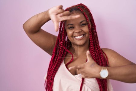Téléchargez les photos : Femme afro-américaine avec des cheveux tressés debout sur fond rose souriant faisant cadre avec les mains et les doigts avec le visage heureux. concept de créativité et photographie. - en image libre de droit
