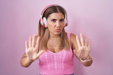 Foto de Mujer rubia joven escuchando música usando auriculares alejando las palmas de las manos mostrando rechazo y negación con miedo y expresión repugnante. parada y prohibido. - Imagen libre de derechos